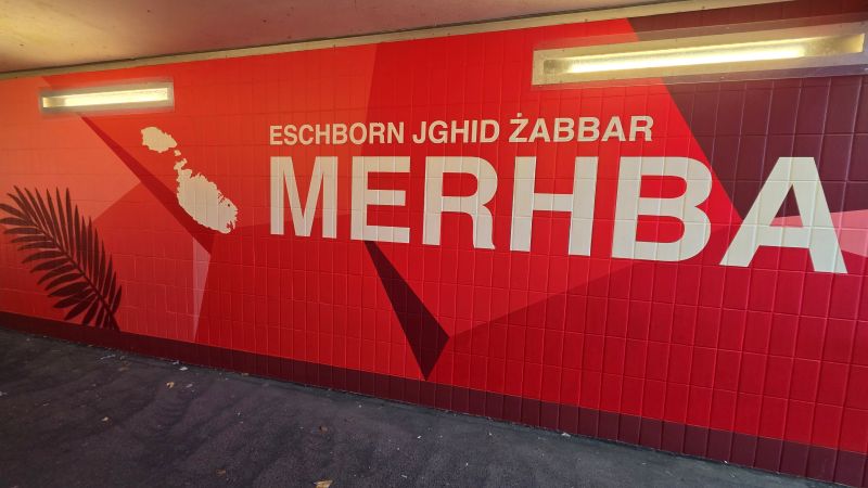 Rotes Wandbild in einer Unterführung mit der maltesischen Begrüßung Eschborn jgħid Żabbar Merħba und den Umrissen von Malta und Gozo.