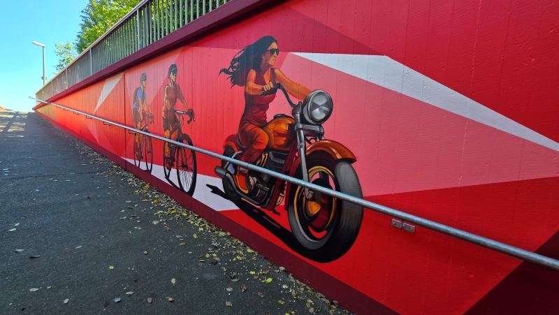 Graffitikunst auf rotem Hintergrund in einer Unterführung, die eine Motorradfahrerin und Radfahrer als Motiv der Pilgerfahrt nach Żabbar zeigt.