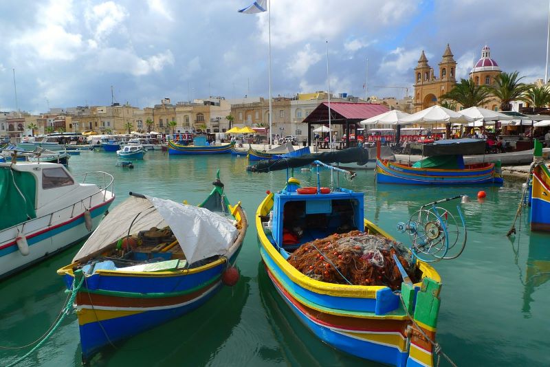 Marsaxlokk mit bunten Fischerbooten im Vorder- und der Kirche Madonna ta' Pompej im Hintergrund. Bild von JanneG auf Pixabay.
